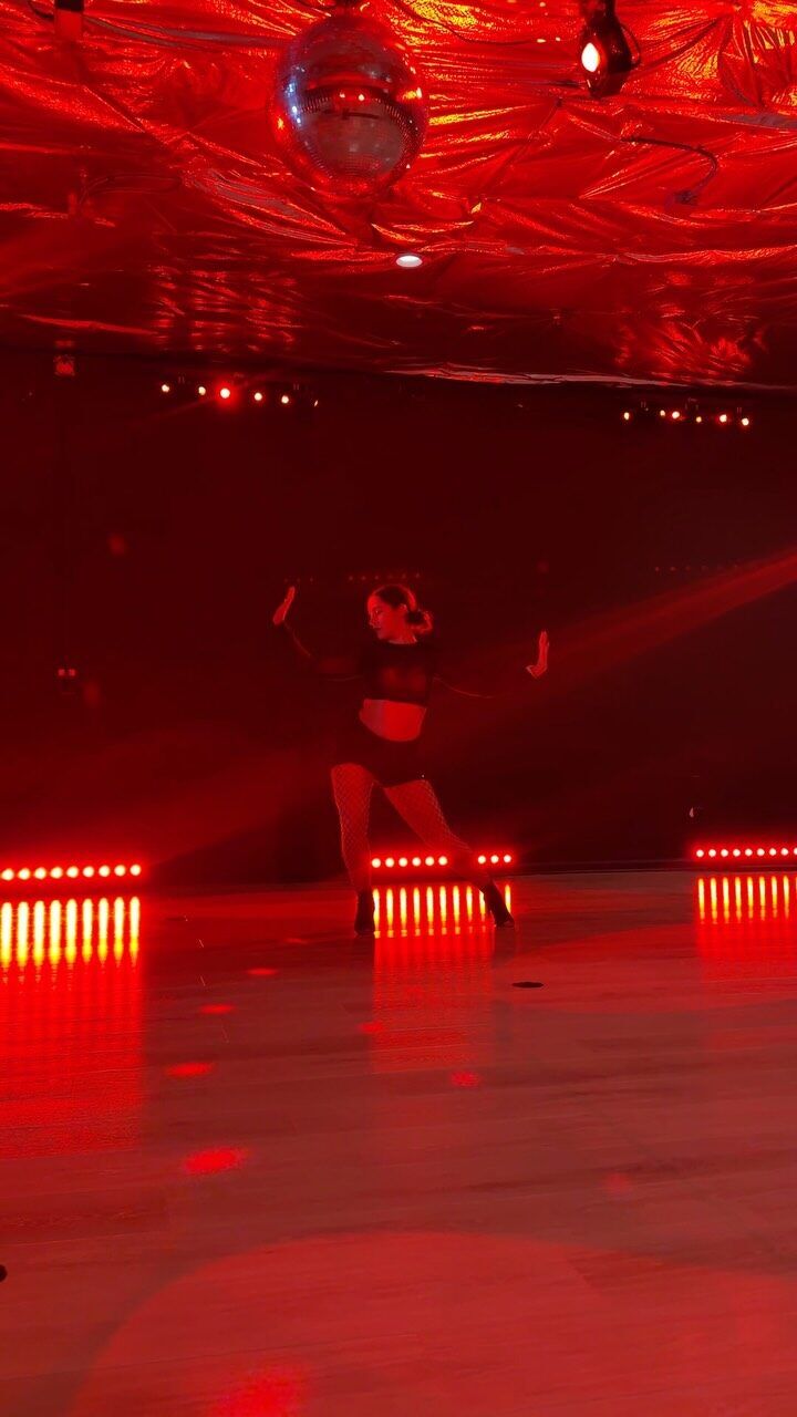 A dancer in fishnets and heels under red light with a disco ball overhead
